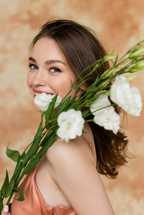 cheerful young woman with brunette hair posing in pink slip dress and holding white eustoma flowers on mottled beige background, sensuality, sophistication, elegance, looking at camera