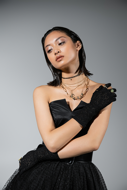 portrait of brunette and asian young woman with short hair posing in black strapless dress and gloves with golden rings, looking away on grey background, necklaces, natural makeup