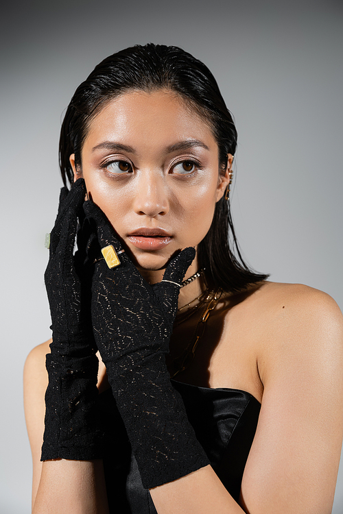 portrait of attractive and asian young woman with short hair posing in black strapless dress and gloves with golden rings, looking away on grey background, wet hairstyle, necklaces, natural makeup