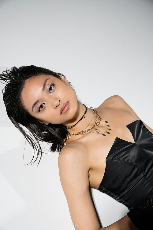 alluring and asian young woman with short hair lying in black strapless dress while posing in golden jewelry on grey background, wet hairstyle, natural makeup, looking at camera