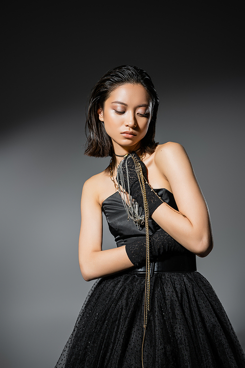 charming asian young woman with short hair holding golden and silver jewelry while wearing gloves and standing in elegant strapless dress on grey background, wet hairstyle, natural makeup