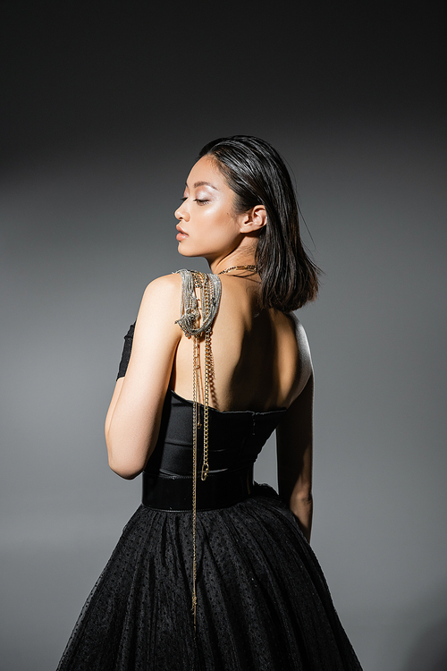 side view of brunette and asian young woman with short hair posing with golden and silver jewelry on shoulder while standing in strapless dress on grey background, wet hairstyle, natural makeup