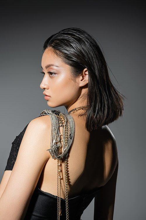 portrait of brunette and asian young woman with short hair posing with golden and silver jewelry on shoulder while standing in strapless dress on grey background, wet hairstyle, natural makeup, side view