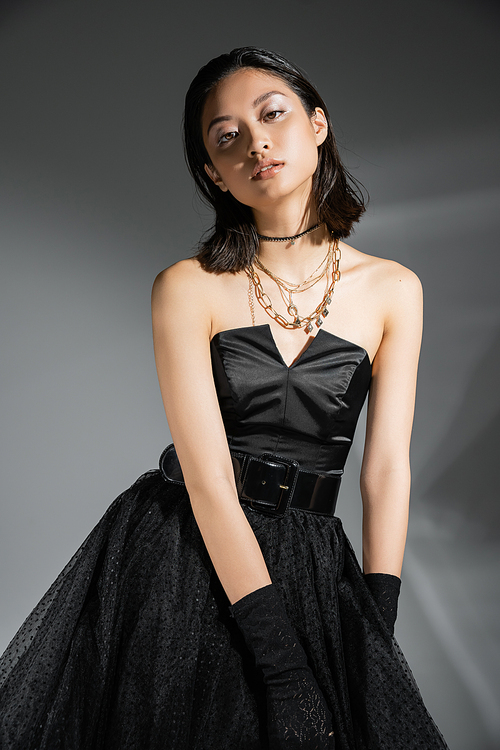 portrait of stylish asian young woman with short hair posing in black strapless dress with belt and gloves while looking at camera on grey background, wet hairstyle, golden necklaces