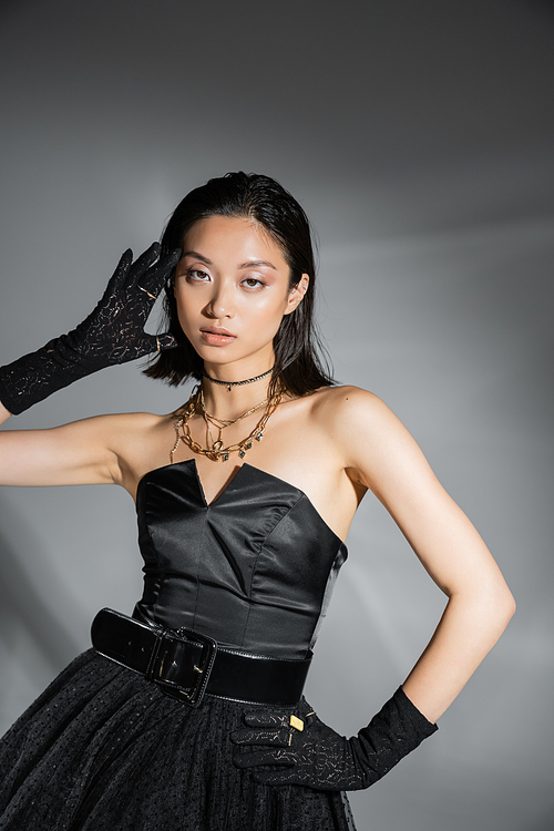 mesmerizing young asian woman with short hair posing with hand on hip in black strapless dress with belt and gloves while looking at camera on grey background, wet hairstyle, golden necklaces