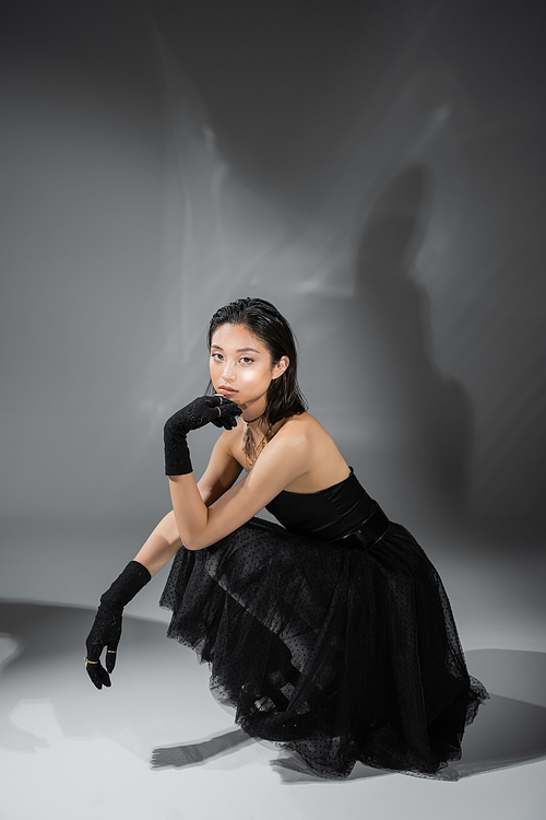 full length of stylish asian young woman with short hair sitting in black strapless dress with tulle skirt and gloves while looking at camera on grey background, wet hairstyle, golden necklaces