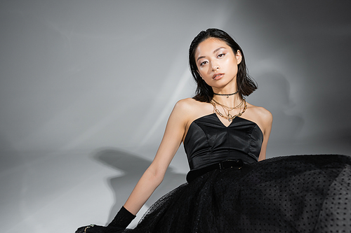 graceful asian woman with short hair sitting in black strapless dress with tulle skirt, belt while looking at camera on grey background, wet hairstyle, golden necklaces, captivating beauty