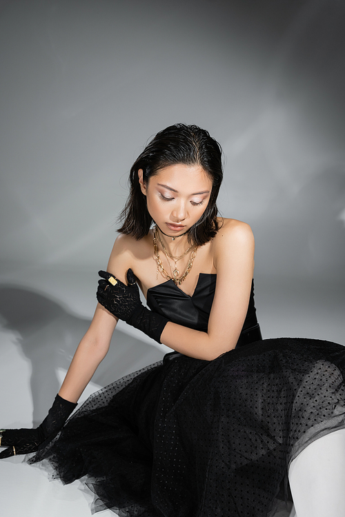 stunning asian young woman with short hair sitting in black strapless dress with tulle skirt and gloves while looking down on grey background, wet hairstyle, golden necklaces and rings