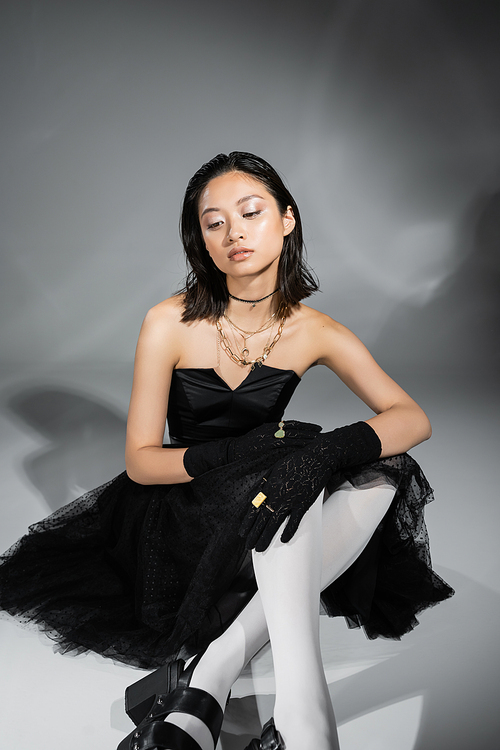 dreamy asian young woman with short hair sitting in black strapless dress with tulle skirt white tights, shoes and gloves while looking away on grey background, wet hairstyle, golden necklaces