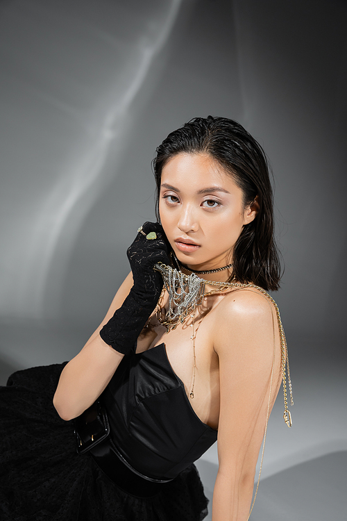 asian model with short and brunette hair holding golden jewelry in hand while posing in strapless dress and black glove on grey background, everyday makeup, wet hairstyle, young woman, glamour