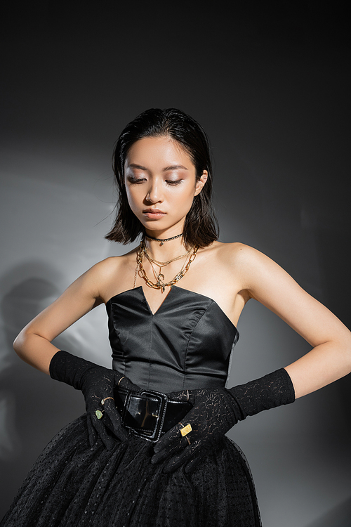 charming asian young woman with short hair posing in black strapless dress with tulle skirt while touching belt and looking down on grey background, wet hairstyle, golden jewelry