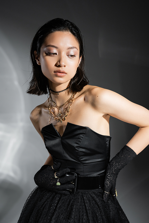 charming asian young woman with short hair posing in black strapless dress with tulle skirt with belt and gloves while looking away on grey background, wet hairstyle, golden jewelry