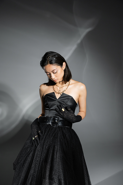 charming asian young woman with short hair posing in black strapless dress with tulle skirt with belt and gloves standing on grey background, wet hairstyle, golden jewelry