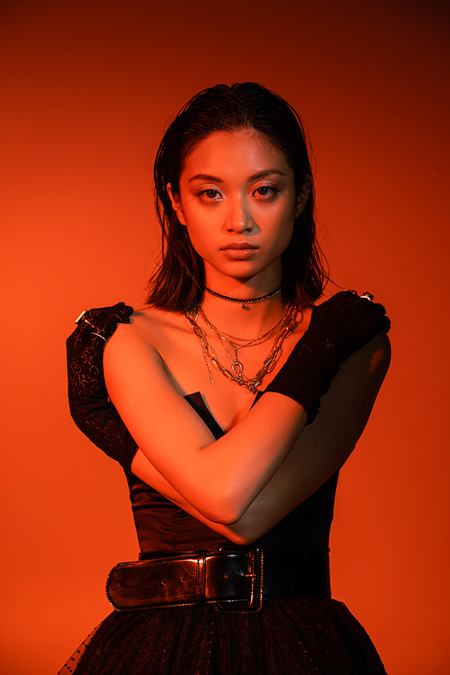 beautiful asian young woman with short hair and wet hairstyle posing with crossed arms in black strapless dress and gloves while standing on orange background with red lighting, golden jewelry