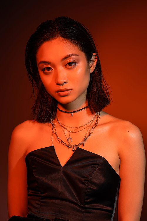 portrait of beautiful asian young woman with short hair and wet hairstyle posing in black strapless dress while standing on orange background with red lighting, golden jewelry