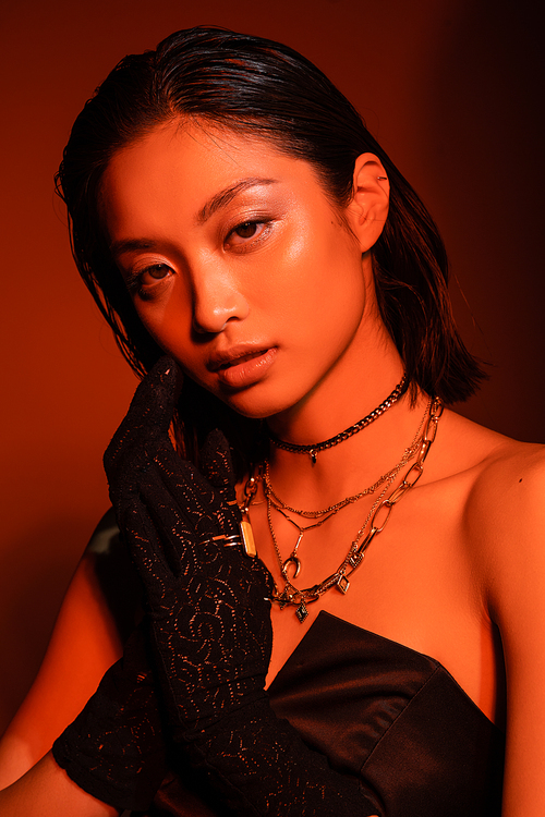 portrait of alluring asian young woman with short hair and wet hairstyle posing in black strapless dress and gloves while standing on orange background with red lighting, golden jewelry