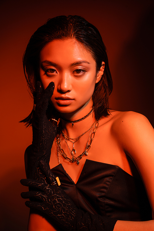 portrait of stunning asian young woman with short hair and wet hairstyle posing in black strapless dress and gloves while standing on orange background with red lighting, golden jewelry