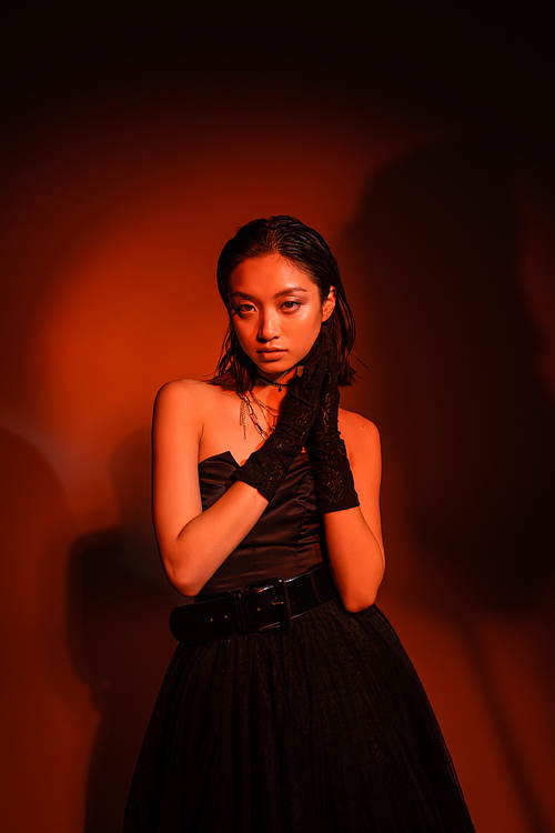 pretty asian woman with short hair and wet hairstyle posing in black strapless dress with tulle skirt and gloves while standing on dark orange background with red lighting, golden jewelry, young model
