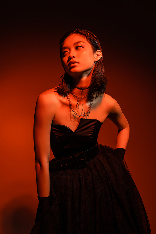 asian woman with short hair and wet hairstyle posing with hand on hip in black strapless dress with tulle skirt and gloves while standing on orange background with red lighting, golden jewelry