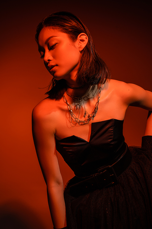 mesmerizing asian woman with short hair and wet hairstyle posing in black strapless dress with tulle skirt and gloves while standing on orange background with red lighting, golden jewelry, young model