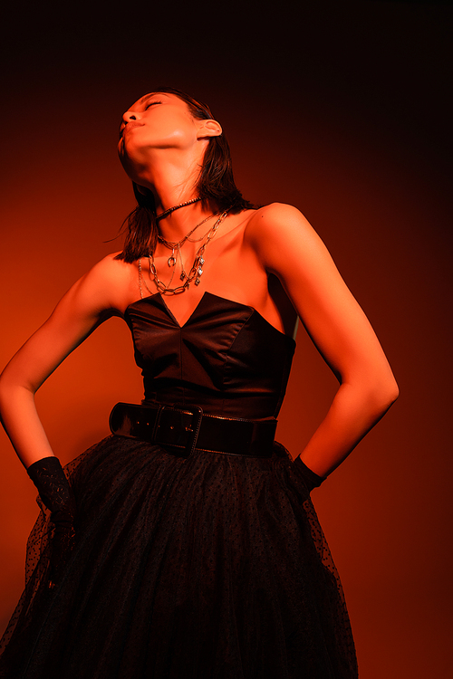 stylish asian woman with closed eyes and wet hairstyle posing in black strapless dress with tulle skirt and gloves while standing on orange background with red lighting, golden jewelry, young model