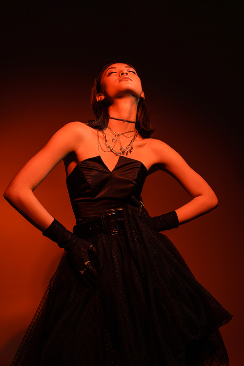 stylish asian woman with closed eyes and wet hairstyle posing with hands on hips in black strapless dress with tulle skirt and gloves while standing on orange background with red lighting, young model