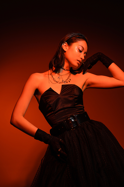 asian woman with short hair and wet hairstyle posing with hand on hip in black strapless dress with tulle skirt and gloves while standing on dark orange background with red lighting, young model