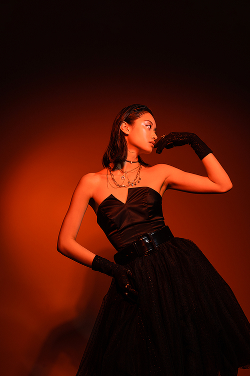 radiant asian woman with short hair and wet hairstyle posing with hand on hip in black strapless dress with tulle skirt and gloves while standing on orange background with red lighting, young model