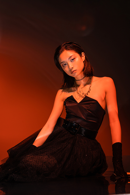 beautiful asian woman with short hair and wet hairstyle posing in strapless dress with tulle skirt and gloves while standing on orange background with red lighting, young model, looking at camera