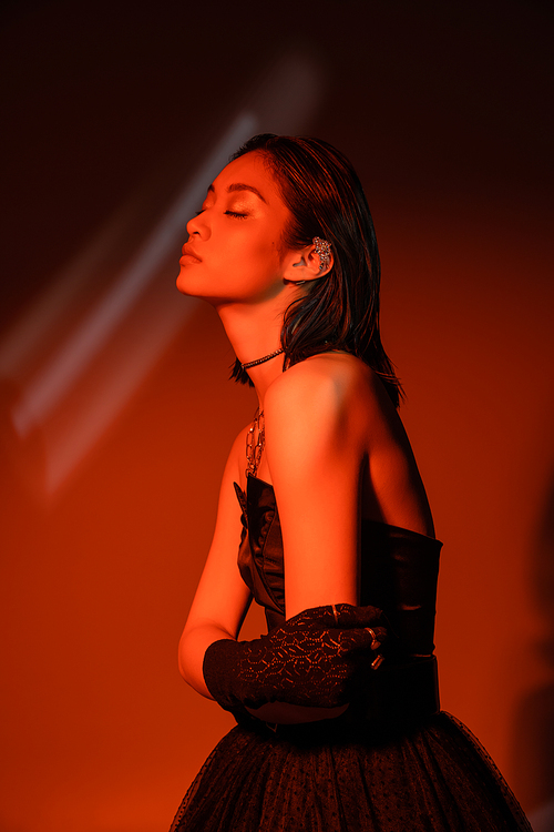 portrait of stunning asian woman with short hair and wet hairstyle posing in strapless dress and glove with trendy cuff earring and necklaces on dark orange background with red lighting, young model