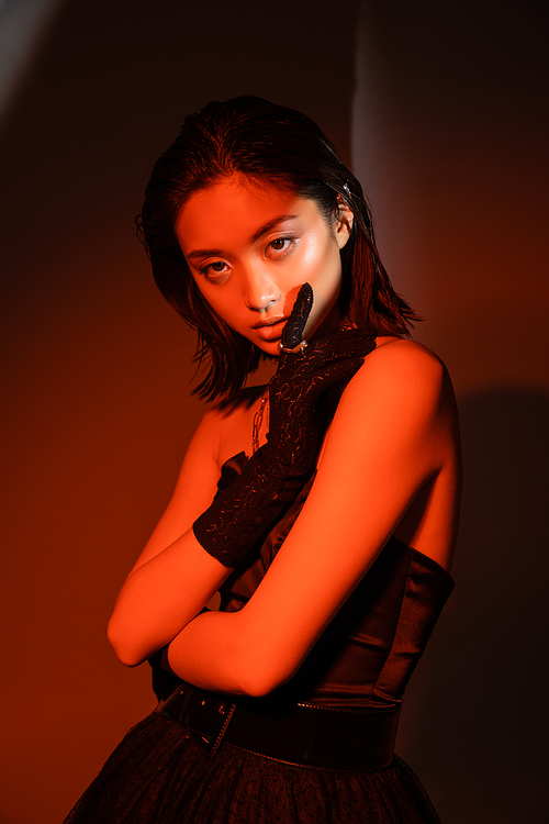 portrait of stunning asian woman with short hair and wet hairstyle posing in strapless dress and glove with golden rings with trendy cuff earring on orange background with red lighting, young model