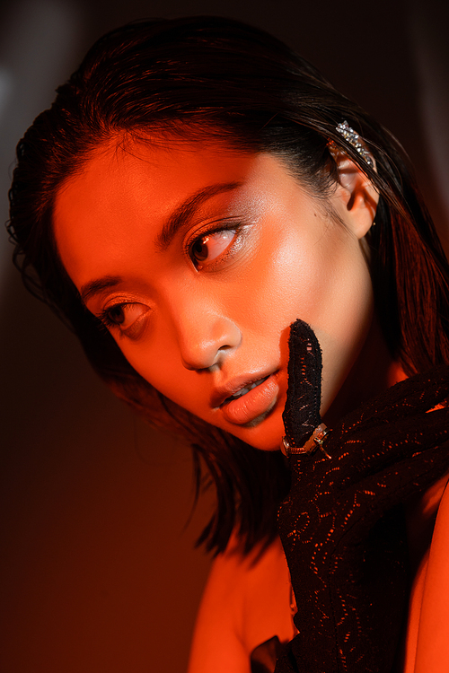 portrait of alluring asian woman with short hair and wet hairstyle posing in black glove with golden rings and looking away on dark background with red lighting, young model, cuff earring