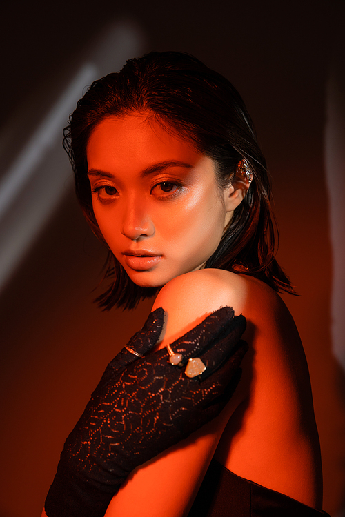 portrait of charming asian woman with short hair and wet hairstyle posing in black glove with golden rings and looking at camera on dark background with red lighting, young model, cuff earring