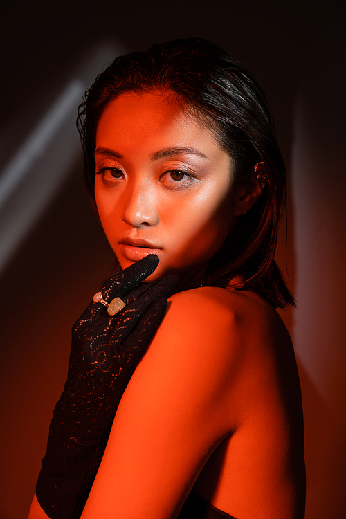 portrait of young asian woman with short hair and wet hairstyle posing in black glove with golden rings and looking at camera on dark background with red lighting, model, cuff earring