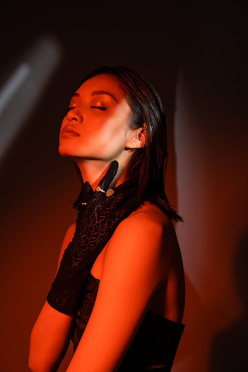 portrait of beautiful asian model with closed eyes and wet hairstyle posing in strapless dress and glove with golden rings and touching neck on dark orange background with red lighting, young woman