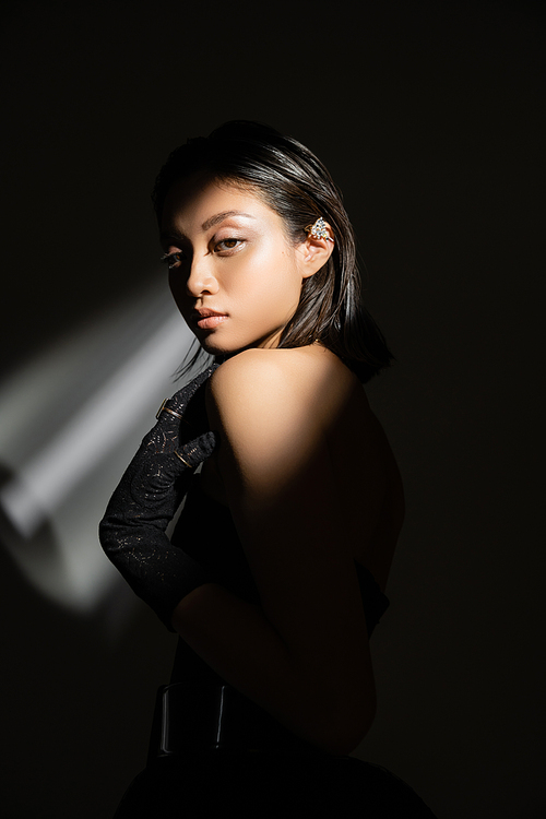 elegant asian young woman with wet hairstyle and short hair posing in strapless dress and black glove while standing on grey background, young model, looking at camera, shadows, dark