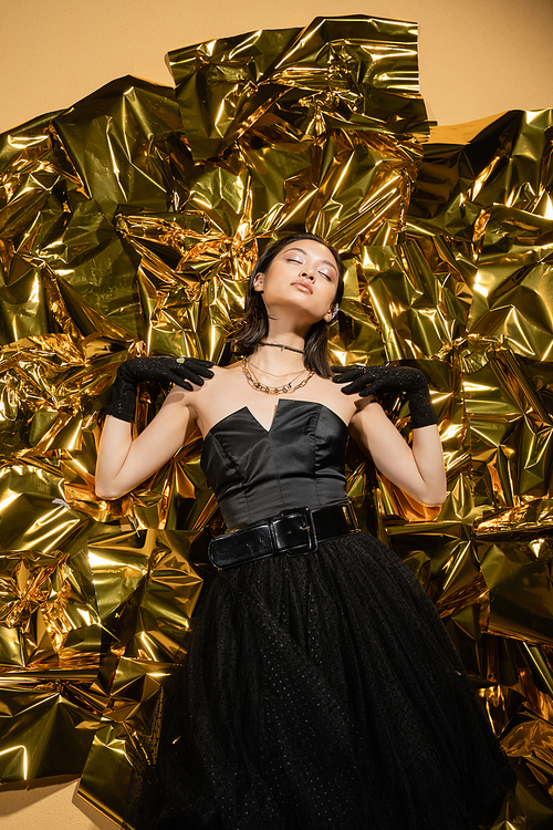elegant asian young woman with short hair and closed eyes posing in black strapless dress with tulle skirt and gloves while standing next to shiny background, model, wrinkled golden foil