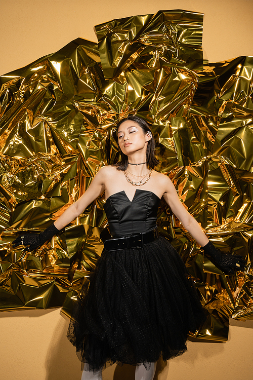 enchanting asian young woman with short hair posing in black strapless dress with tulle skirt and gloves while standing next to shiny yellow background, model, wrinkled golden foil, looking away