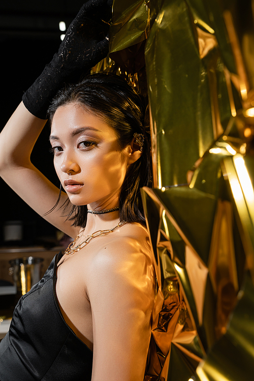 asian young woman with wet short hair posing in strapless dress with black glove and ear cuff while standing next to shiny background, model, looking at camera, wrinkled golden foil