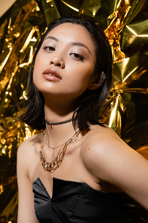 beautiful asian young woman with short hair posing in strapless dress with black glove while standing next to shiny yellow background, model, looking at camera, wrinkled golden foil, wet hairstyle