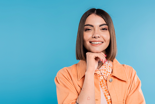 summer fashion, cheerful young woman with short hair and piercing in nose and tattoos looking at camera on blue background, everyday makeup, orange shirt, generation z