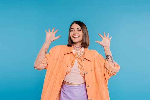 excited young woman gesturing and smiling while looking at camera, on blue background, summer outfit, generation z, short brunette hair, orange shirt, pierced nose, tattooed