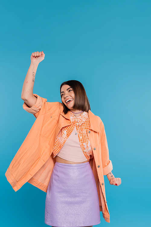 summer outfit, excited young woman smiling while gesturing on blue background, summer outfit, generation z, short brunette hair, orange shirt, pierced nose, tattooed