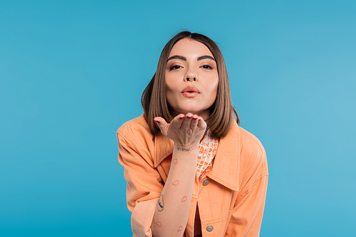 sending air kiss, brunette young woman with short hair, piercing in nose and tattoos posing in casual outfit on blue background, everyday makeup, orange shirt, generation z, blow kiss