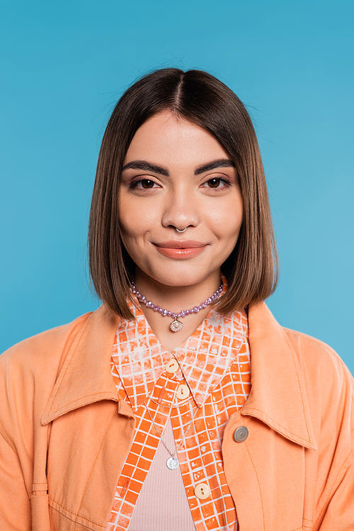 portrait of cheerful woman, young fashion model smiling and looking at camera on blue background, orange shirt, generation z, short brunette hair, pierced nose, summer outfit, gen z fashion
