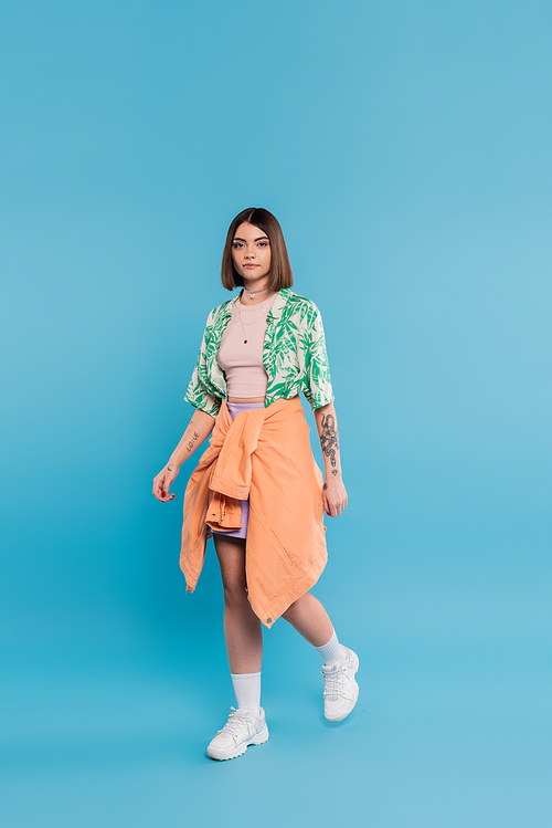 everyday style, young brunette woman with short hair walking in shirt with palm tree print, skirt and white sneakers on blue background, carefree, tattooed, nose piercing, casual attire, full length