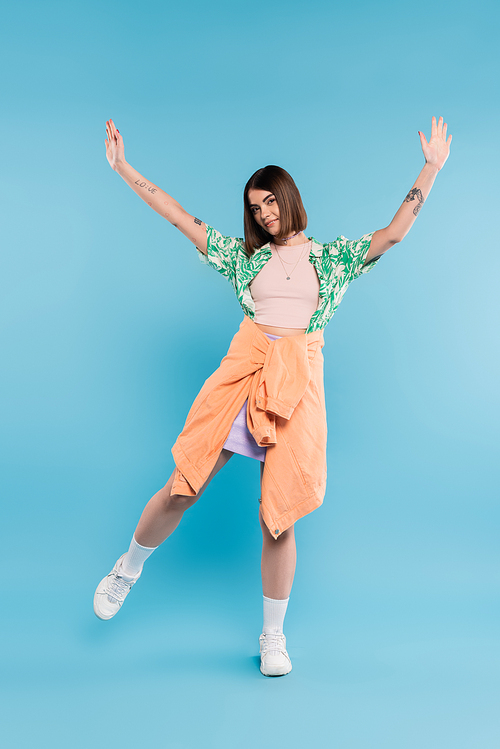 gen z fashion, carefree woman with short hair posing in shirt with palm tree print, skirt and white sneakers on blue background, tattooed, nose piercing, casual attire, raised hands, full length