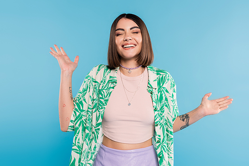 tattooed young woman with short brunette hair wearing shirt with palm tree print, smiling with closed eyes and gesturing with hands on blue background, casual attire, gen z fashion, happiness