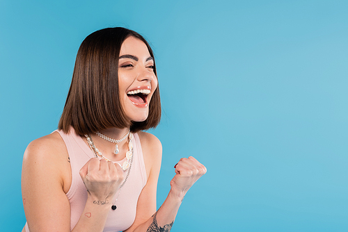 excitement, tattooed young woman with short brunette hair in tank top smiling and gesturing with hands on blue background, casual attire, gen z fashion, happiness