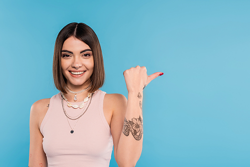 cheerfulness, tattooed young woman with short brunette hair in tank top smiling and pointing with thumb on blue background, casual attire, gen z fashion, happiness, showing something on camera